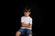 © Corina - A little boy sitting on chair, hands crossed, being upset, on black background.