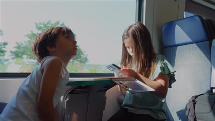 Wall Mural - Two children sitting together on a train, engaged in an activity with books, focused on reading and writing during a daytime journey through a scenic landscape