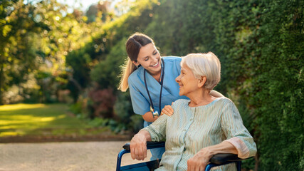 Happy nurse caring for elderly woman in a wheelchair. Young female pushing senior retirement nursing home patient in a wheelchair outdoors in a garden