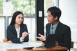 © Satori Studio - Two business professionals engaged in a discussion during a meeting in a modern office environment, with documents and a laptop on the table.