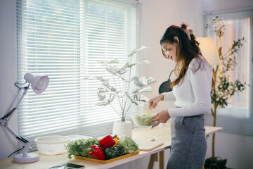 Young woman is preparing a healthy vegan meal, adding fresh lettuce to a glass bowl in a modern kitchen with natural light