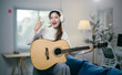 © Parichat - Young woman is playing acoustic guitar in her living room and enjoying music. She is wearing headphones and making rock sign with hand while sitting on the sofa