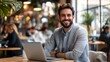 © Pattanan - A smiling businessman with a laptop, sitting at a desk in a coworking space, surrounded by bustling activity