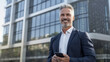 © halo - A businessman holding a mobile phone in front of an office building, smiling at the camera while standing outdoors near a skyscraper.