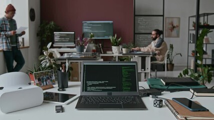 Timelapse of laptop on desk in IT company office, lines of computer code running on black screen, VR headset. Team of multiethnic male, female programmers working together on project in background