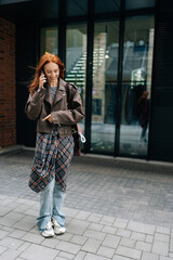 Wall Mural - Full length vertical portrait of attractive young woman with flowing red hair enjoying cheerful phone conversation standing on city street on summer day. Smiling redhead female contemporary lifestyle