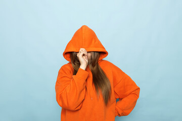 Young woman in orange hoodie on blue background hides face under hood