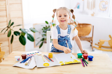 a little child girl paints a drawing with a brush in an album and smiles, a happy child draws a gouache picture at home at the table