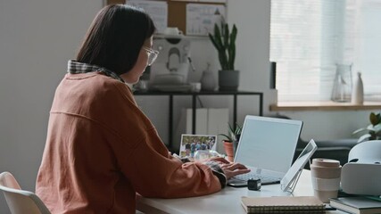 Medium rear shot of young Asian female programmer in orange sweatshirt sitting at desk, writing computer software code on laptop and checking it on tablet while working alone in office