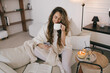 © polinaloves - Young woman using her smartphone and drinking tea, relaxing in a cozy living room.