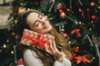 © polinaloves - Young woman holding Christmas present with a red ribbon, standing near Christmas tree.