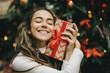 © polinaloves - Young woman holding Christmas present with a red ribbon, standing near Christmas tree.