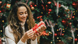 © polinaloves - Young woman holding Christmas present with a red ribbon, standing near Christmas tree.