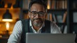 © QITIAN - Smiling middle-aged man with glasses working on a laptop in a cozy home office.