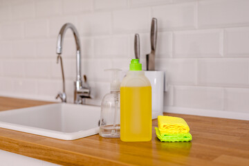 White plates, glass cups and forks in a clean white sink and two bottles of detergent and microfiber rags on a wooden table in a white kitchen