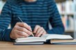 © Vilaysack - Close-up of a person holding a pencil, writing notes in an open book, symbolizing learning, studying, or journaling in a focused setting.