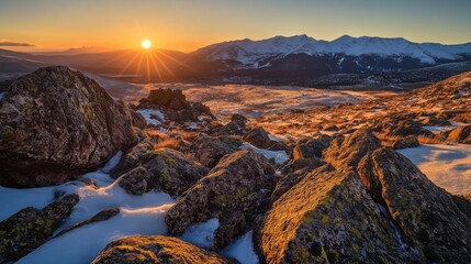 Poster - A dramatic sunrise scene with the sun peeking over a distant mountain ridge, casting long shadows and warm light across a rugged landscape of rocks and snow.