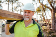 © Austockphoto - A man wearing hardhat carrying a stack of lumber on his shoulder at a construction site.
