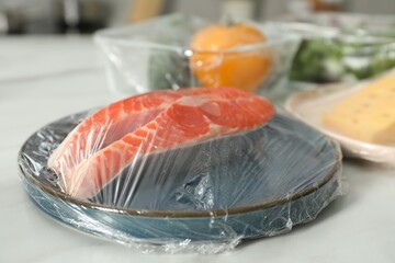 Plate of salmon with plastic food wrap on white table, closeup