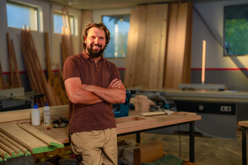 A portrait of smiling carpenter who stands with crossed hands in a woodworking shop, dressed in working suit, surrounded by stacks of lumber and leaned on working table.