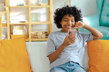 Wall Mural - Excited happy young black african american woman holding cell phone laughing feeling joy getting mobile message. Overjoyed girl laughing aloud sitting on couch watching funny video reading news