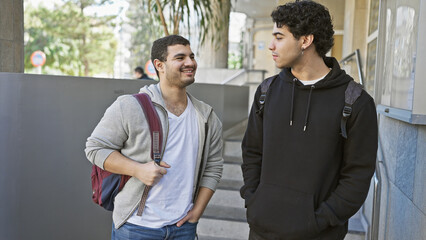 Two young men, possibly friends, chat on a city street with an urban backdrop.