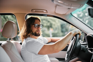 Wall Mural - Young man driving a car, looking confidently through the window, with a casual outfit and sunglasses, set in a sunny environment