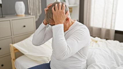 Canvas Print - A distressed grey-haired man sitting on a bed in a bedroom, portraying a scene of worry or headache.