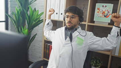 Poster - A young arab man in a white lab coat stretching at his desk in a clinic office, wearing headphones.