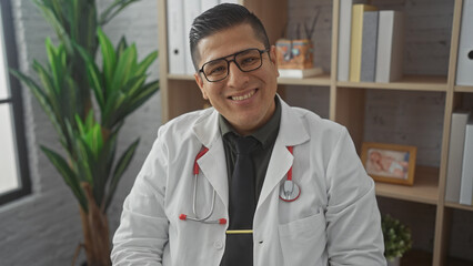 Canvas Print - A smiling hispanic man in a white lab coat with a stethoscope poses confidently in a clinic office.