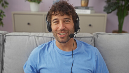 Wall Mural - A handsome young man wearing a headset smiles while sitting on a couch indoors, exuding casual professionalism.