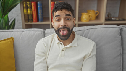 Sticker - A playful young hispanic man sticking his tongue out while relaxing on a sofa in a cozy living room.