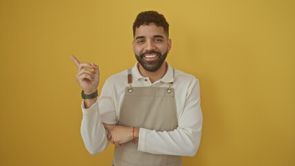 Sticker - Handsome hispanic man smiling and pointing in apron over yellow background