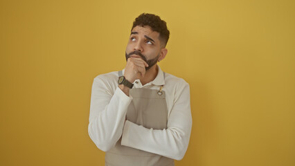 Sticker - Pensive hispanic man with beard wearing apron over isolated yellow background, thinking.