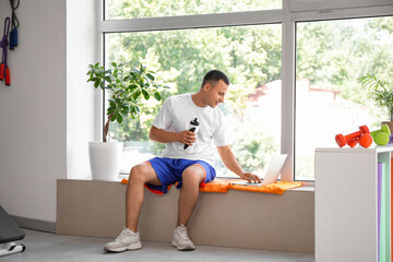 Poster - Sporty young man with water bottle using laptop in gym