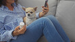 © Krakenimages.com - A middle-aged woman sitting indoors with a chihuahua and holding a pill bottle and cellphone in a casual home setting.