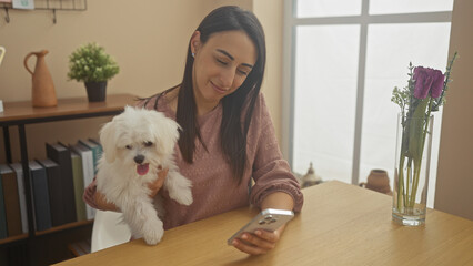 Wall Mural - A smiling young woman holds a bichon maltese dog while looking at a smartphone in a cozy living room.