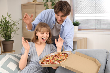 Wall Mural - Young man holding cardboard box with tasty pizza and his surprised girlfriend sitting on sofa in living room