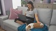 © Krakenimages.com - A smiling young woman works on her laptop from the comfort of her living room couch, accompanied by her sleeping chihuahua.