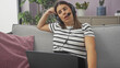 © Krakenimages.com - A young brunette woman wearing a headset looks tired while working on her laptop in a modern living room.
