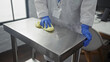 © Krakenimages.com - A woman wearing blue gloves cleans a stainless steel table in a clinic or hospital room with a yellow cloth, indicating a sanitary indoor workplace environment.