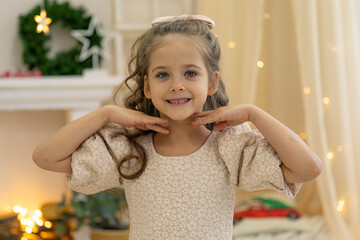 Portrait of cute smiling little toddler girl child on festive bright Christmas decorations background. Looking at camera. Hands near her face