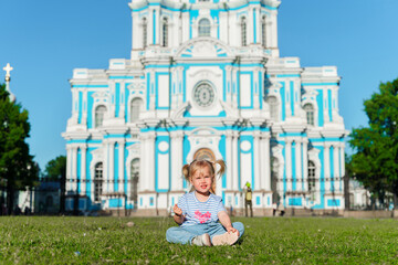 Wall Mural - Little child girl sits on the grass in front of Smolny Cathedral in St. Petersburg on a sunny summer day. The concept of active childhood and tourism.