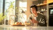 © Prostock-studio - A young man is seated at a kitchen table, eating a meal while looking at his phone. He is wearing headphones and is sitting in front of a window with sunlight streaming in.