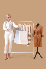 Sticker - Happy mature female stylist holding shopping bags near rack with stylish clothes and mannequin on beige background