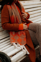 Wall Mural - Fashion cloth details of warm brown coat, cozy pants and orange scarf, woman sitting on bench in autumn park