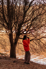 Wall Mural - Brunette woman in autumn bright orange sweater drinking coffee and walking in autumn park by the lake