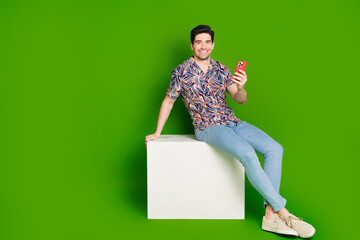 Poster - Full size photo of cheerful guy dressed print shirt jeans sit on white platform holding smartphone isolated on green color background