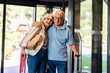 © lordn - An elderly couple enters a hotel lobby, pulling their suitcases. They are dressed casually, smiling, and ready for travel, suggesting they are on a vacation or trip.