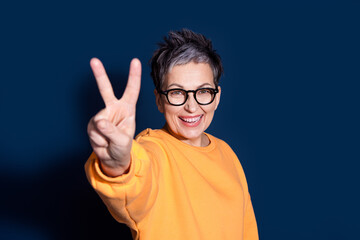 Wall Mural - Photo portrait of pretty senior female show v-sign wear trendy yellow outfit isolated on dark blue color background